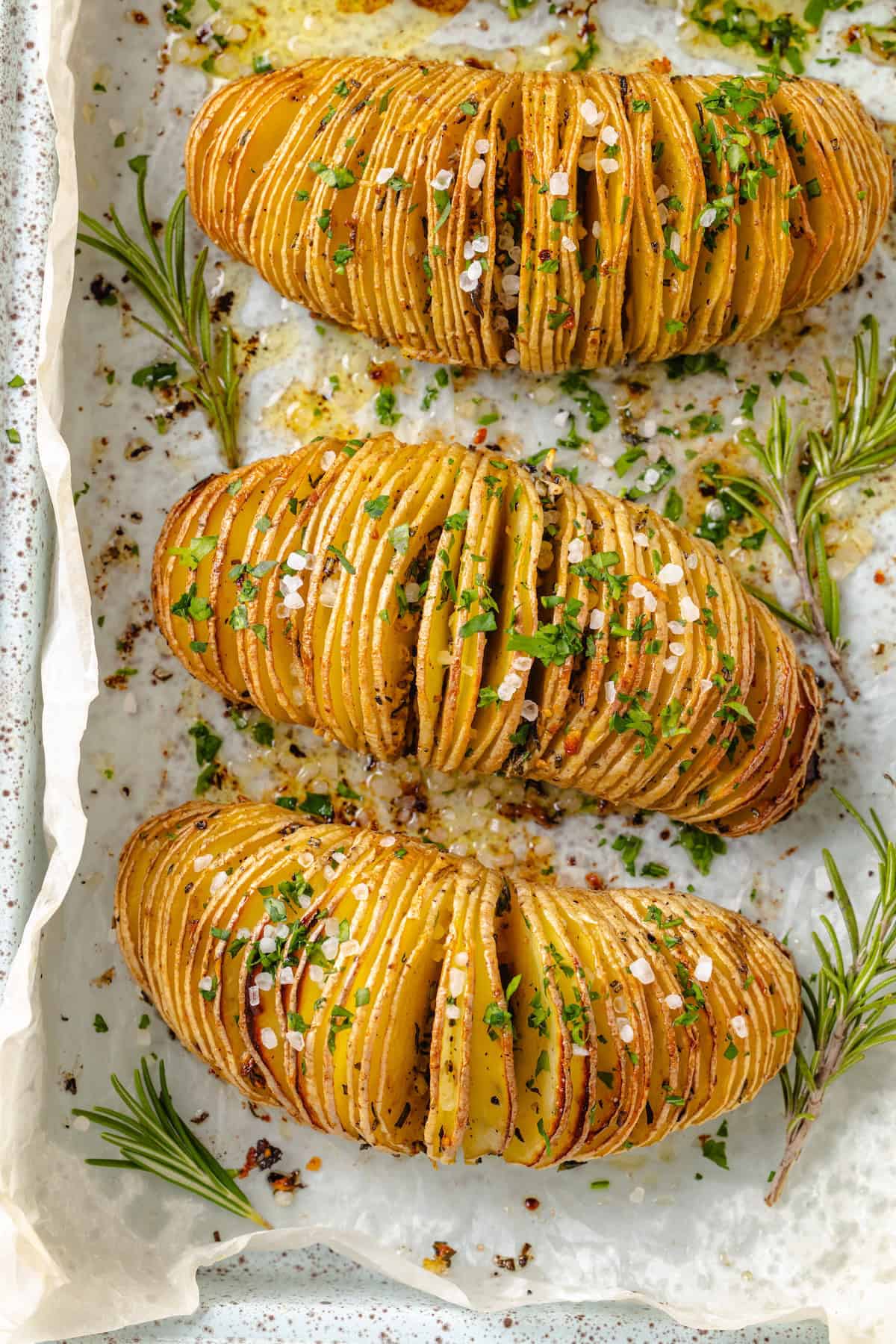 Hasselback potatoes are perfect for a party! The edges get crispy, the insides are tender, and garlic herb butter makes them irresistible. 3 hasselback potatoes on sheet pan with herbs and flaky salt.