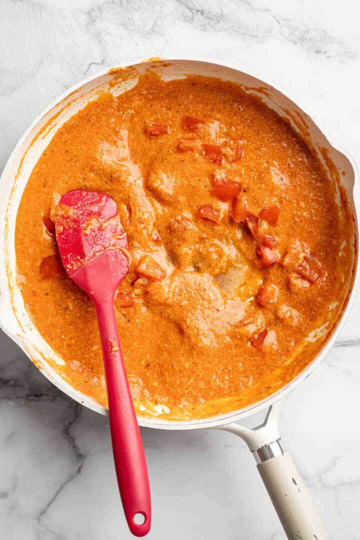 Stirring tomato sauce and diced tomatoes into a skillet with plant milk and melted vegan mozzarella.