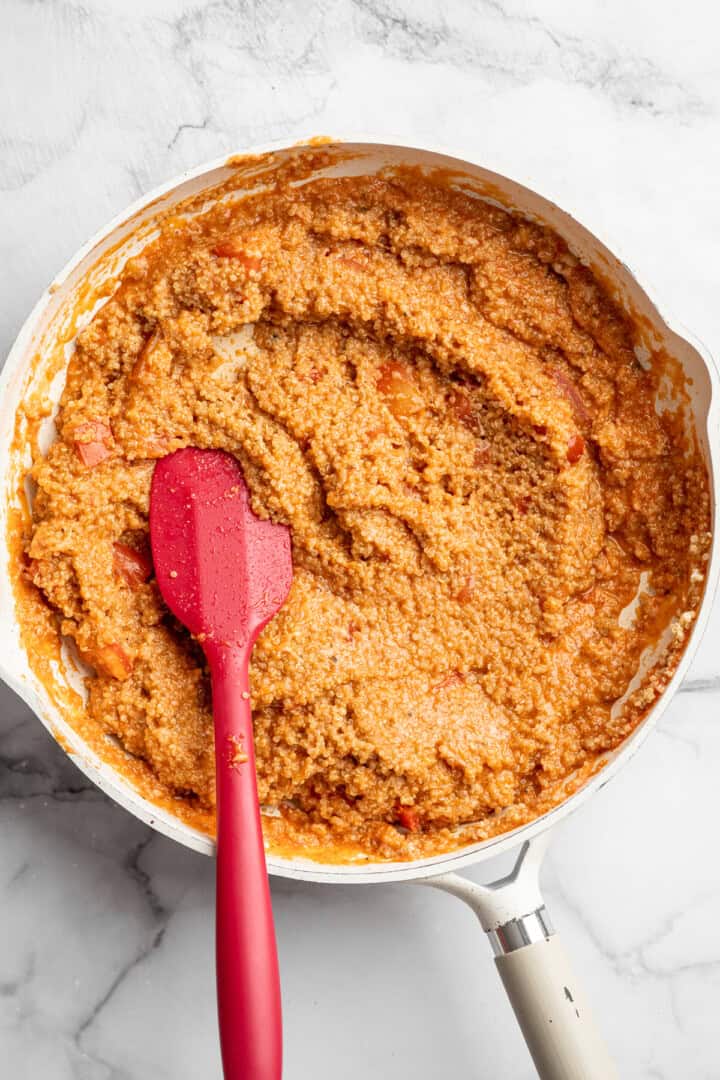 Stirring cooked quinoa into a mixture of tomato sauce, diced tomatoes, plant milk, and melted vegan cheese.