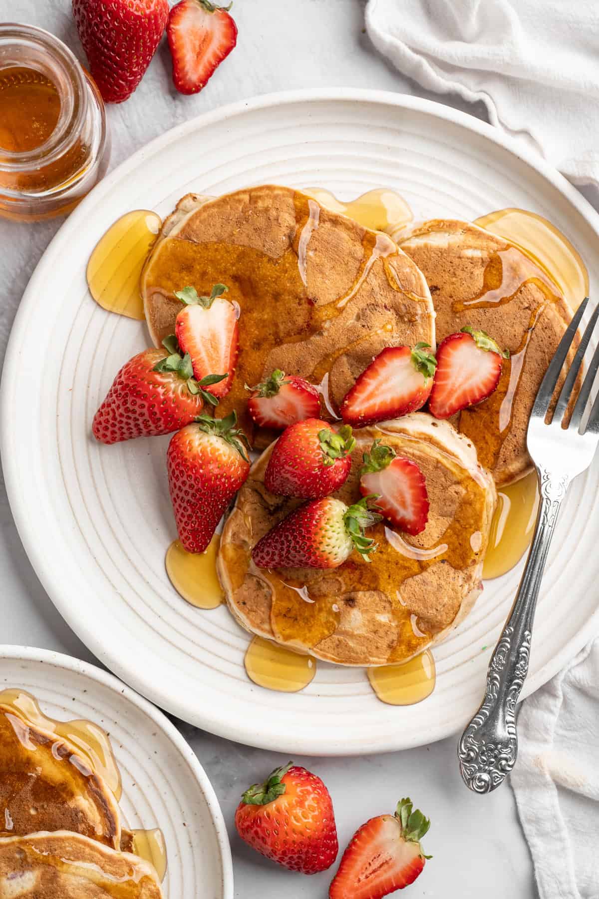 These strawberry pancakes are incredibly fluffy and jam-packed with fresh strawberries. Plus, they're gluten-free and vegan! Overhead image of a plate loaded with strawberry pancakes topped with maple syrup and fresh strawberries.