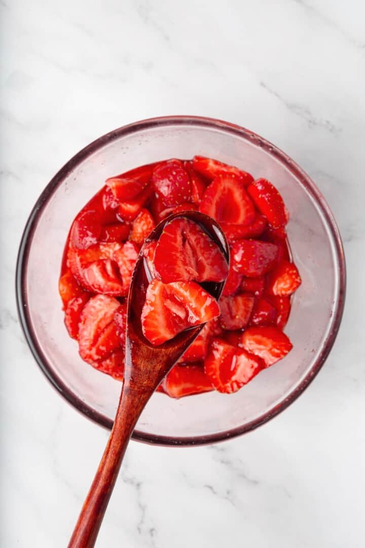 These beautifully simple macerated strawberries are so sweet and syrupy. They're a perfect addition to the breakfast OR dessert table. A wooden spoon holding some macerated strawberries up to the cameral.