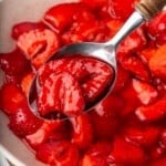 A spoon holding some macerated strawberries above a bowl full of them.