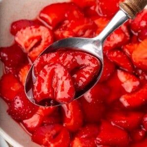 A spoon holding some macerated strawberries above a bowl full of them.