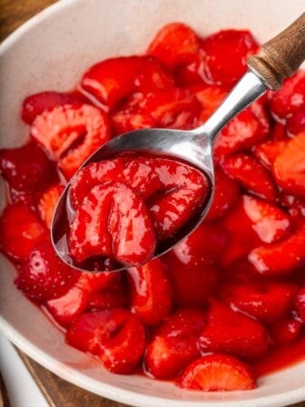 A spoon holding some macerated strawberries above a bowl full of them.