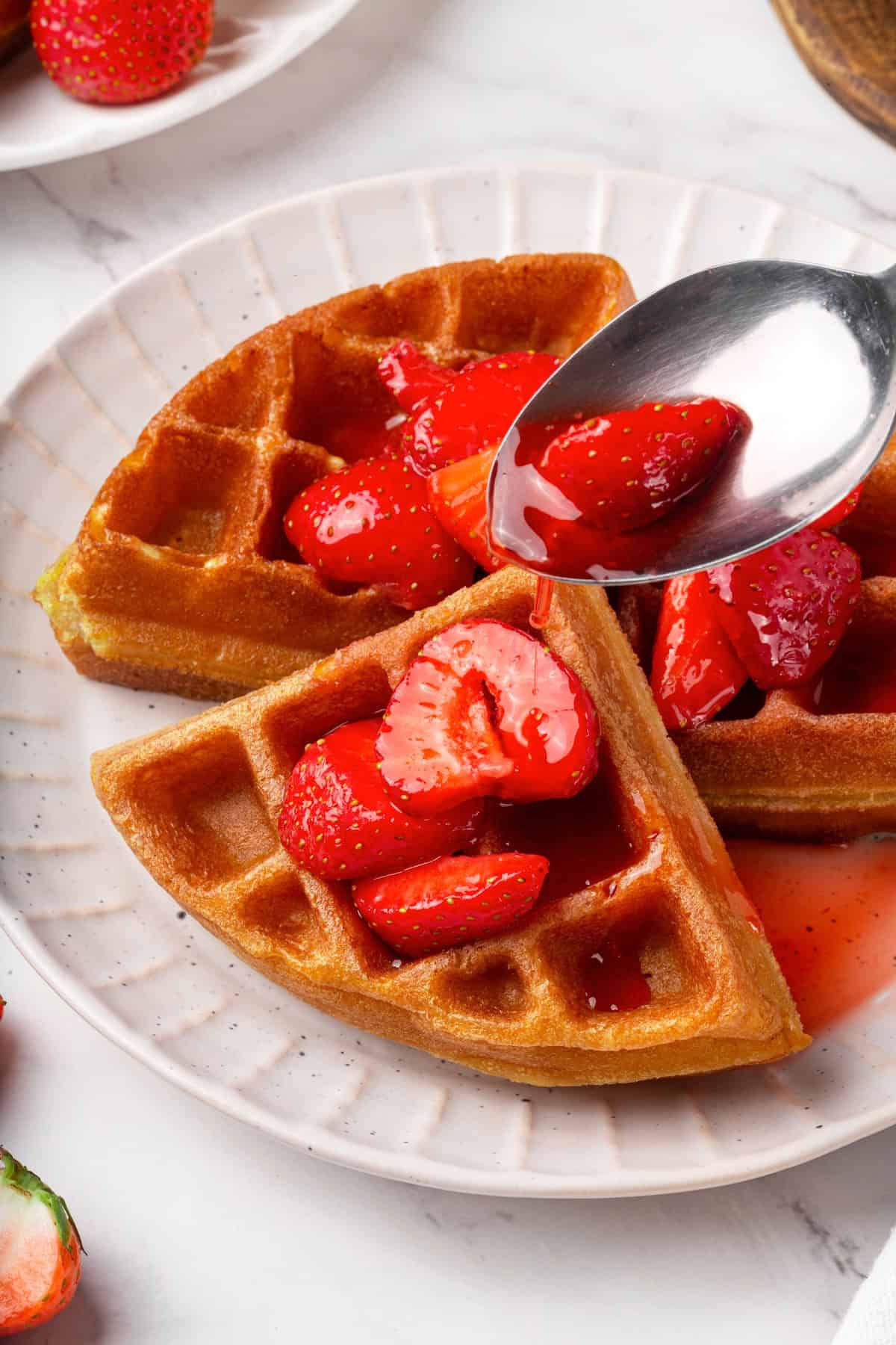 These beautifully simple macerated strawberries are so sweet and syrupy. They're a perfect addition to the breakfast OR dessert table. Close-up shot of a plate of waffles topped with macerated strawberries. A spoon holds a berry up to the camera.