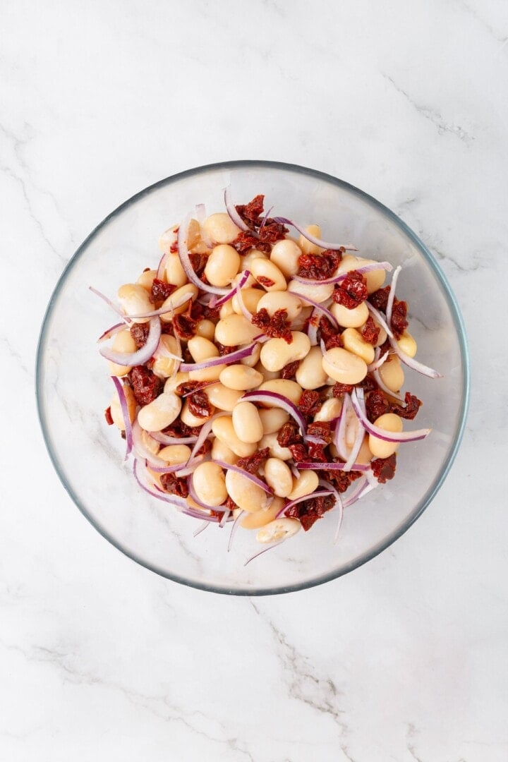 These easy marinated beans are soaked in a bright lemon-Dijon marinade with fresh herbs and aromatics. The perfect summer side. White beans, sliced red onions, and sun-dried tomatoes in a bowl.