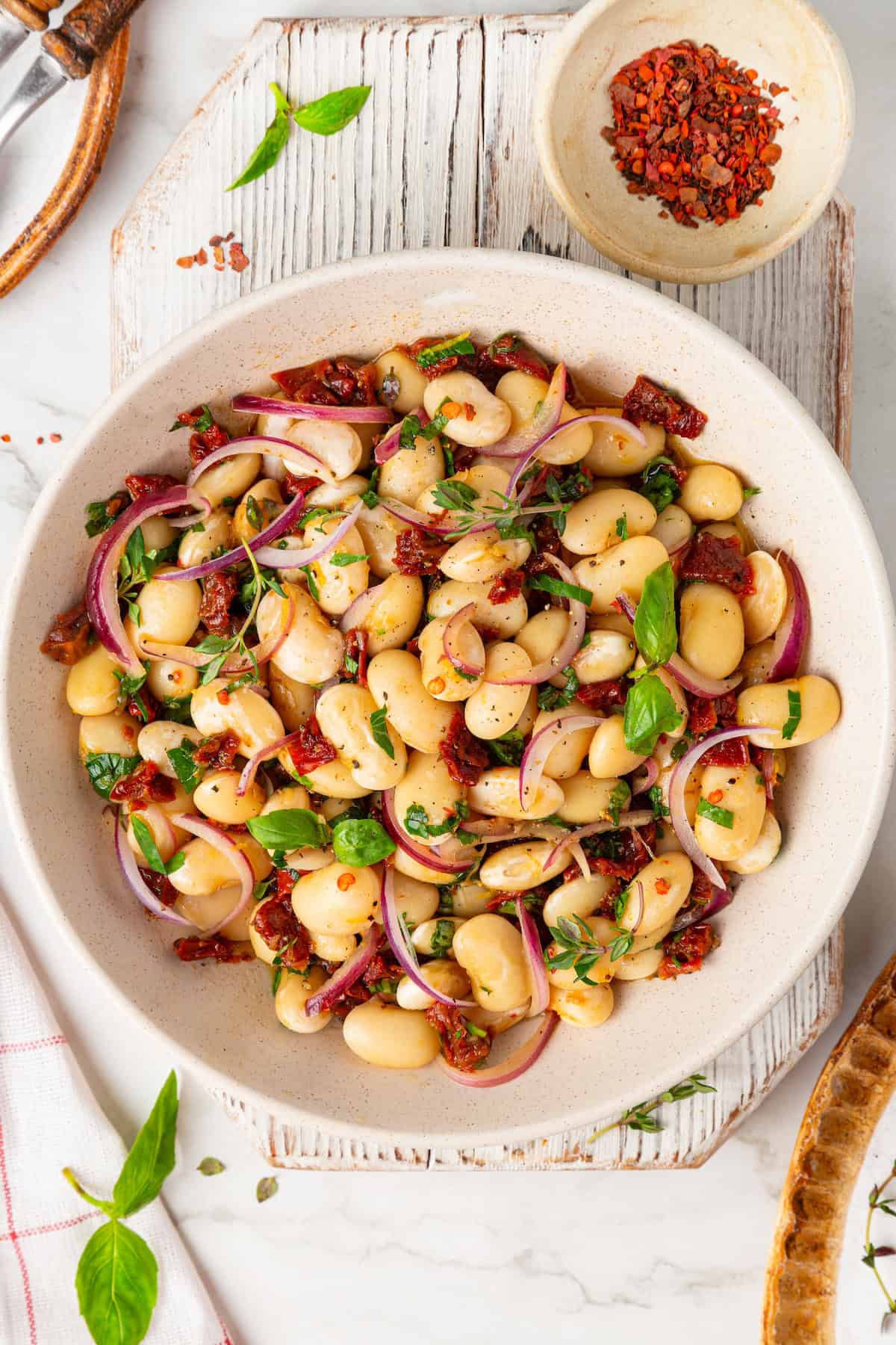 These easy marinated beans are soaked in a bright lemon-Dijon marinade with fresh herbs and aromatics. The perfect summer side. Overhead image of marinated white beans in a serving bowl near a small bowl of red chili flakes.