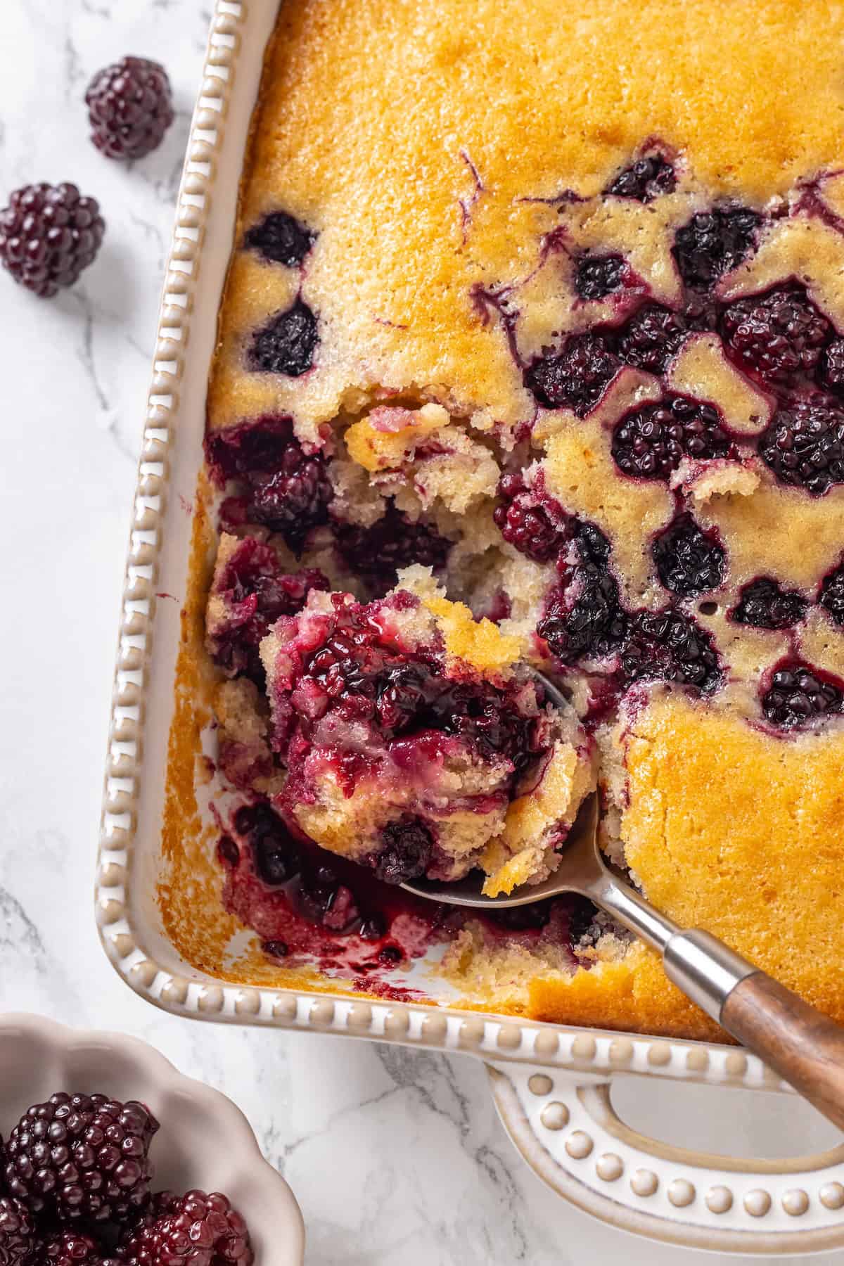 This blackberry cobbler, with its fluffy batter topping and juicy, bubbling blackberries is a summer staple you don't want to miss. Overhead image of a spoon diving into a baking pan full of blackberry cobbler.