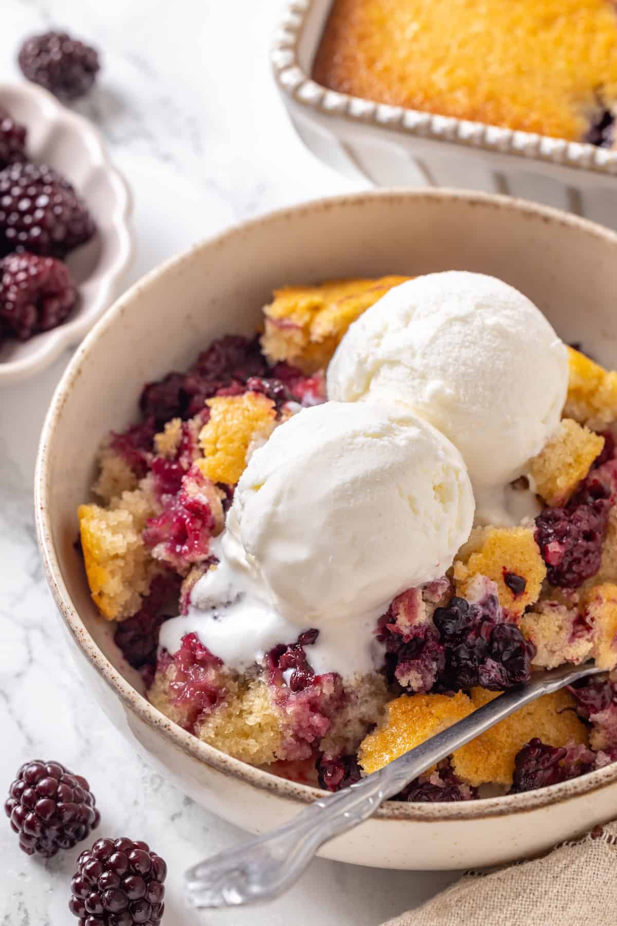 This blackberry cobbler, with its fluffy batter topping and juicy, bubbling blackberries is a summer staple you don't want to miss. Blackberry cobbler topped with vanilla ice cream in a bowl.