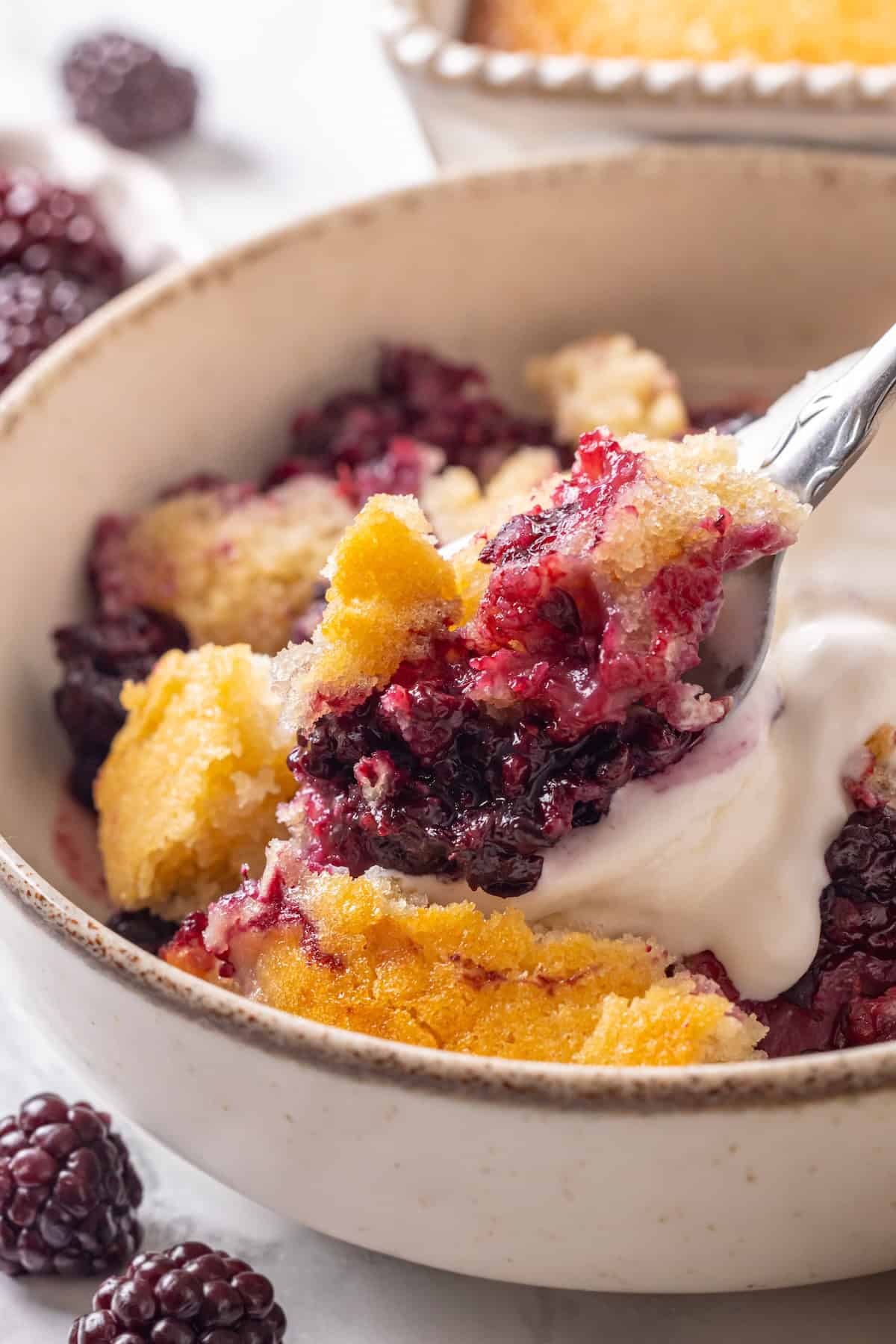 This blackberry cobbler, with its fluffy batter topping and juicy, bubbling blackberries is a summer staple you don't want to miss. Close-up image of a spoon scooping up a bite of blackberry cobbler.