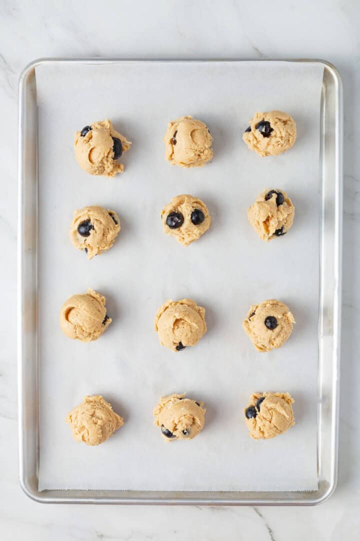 Loaded with jammy pockets of fresh berries and topped with a sweet lemon glaze, these vegan blueberry cookies are a summer must. Balls of lemon blueberry cookie dough arranged on a cookie sheet.