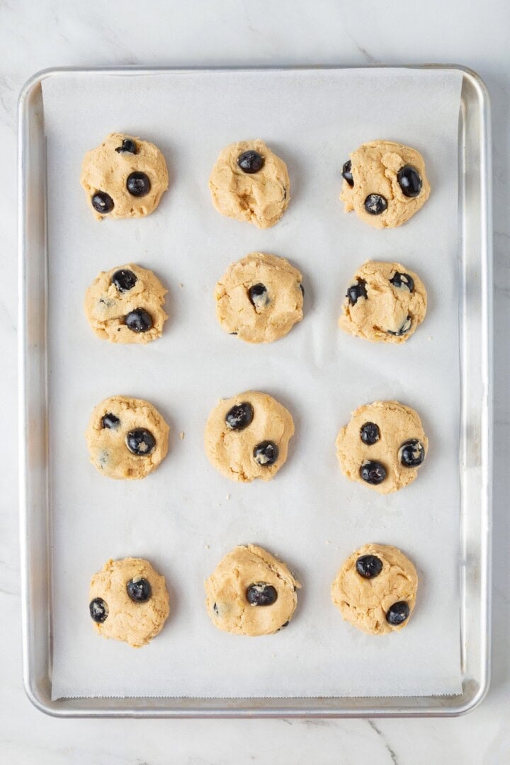 Loaded with jammy pockets of fresh berries and topped with a sweet lemon glaze, these vegan blueberry cookies are a summer must. Balls of lemon blueberry cookie dough shaped into thick disks on a cookie sheet.