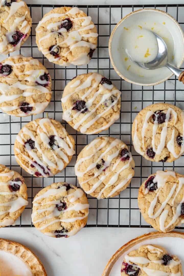 Loaded with jammy pockets of fresh berries and topped with a sweet lemon glaze, these vegan blueberry cookies are a summer must. Overhead image of glazed blueberry cookies on a cooling rack with a bowl of lemon glaze.