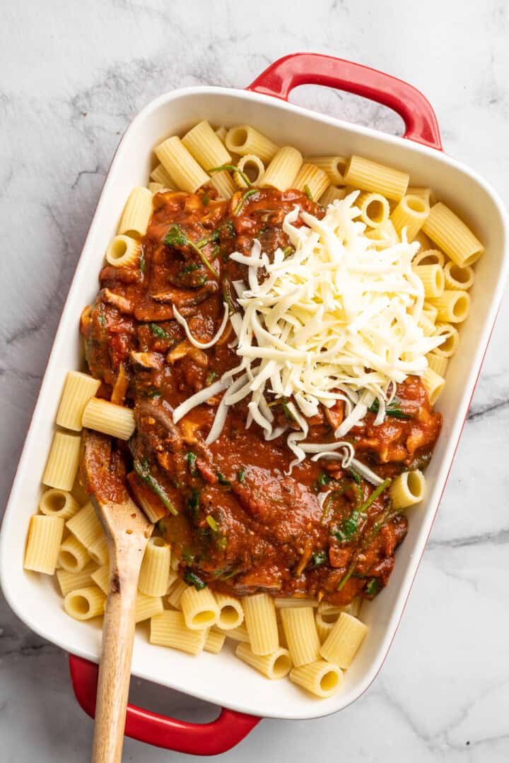 Saucy and loaded with veggies, this vegan cheesy pasta bake is topped with bubbly, melty vegan cheese. It's the ideal weeknight comfort food. Adding veggie-loaded tomato sauce and vegan cheese to a casserole dish with cooked rigatoni.