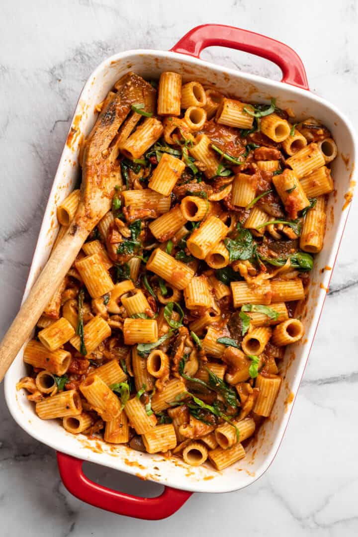 Saucy and loaded with veggies, this vegan cheesy pasta bake is topped with bubbly, melty vegan cheese. It's the ideal weeknight comfort food. Cooked rigatoni in a casserole dish, coated with veggie-loaded tomato sauce.