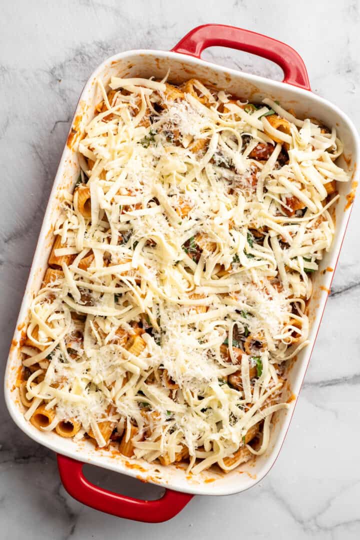 Saucy and loaded with veggies, this vegan cheesy pasta bake is topped with bubbly, melty vegan cheese. It's the ideal weeknight comfort food. Adding vegan mozzarella and parmesan to the top of saucy cooked rigatoni.