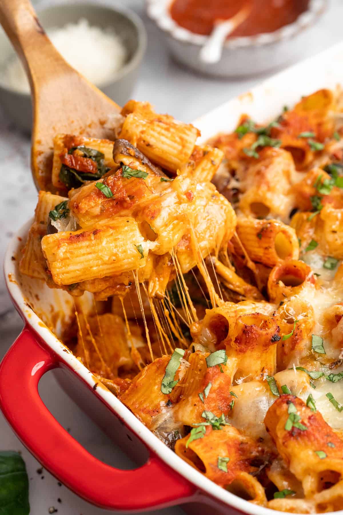 Saucy and loaded with veggies, this vegan cheesy pasta bake is topped with bubbly, melty vegan cheese. It's the ideal weeknight comfort food. A wooden spatula scooping up a serving of baked vegan cheesy pasta.