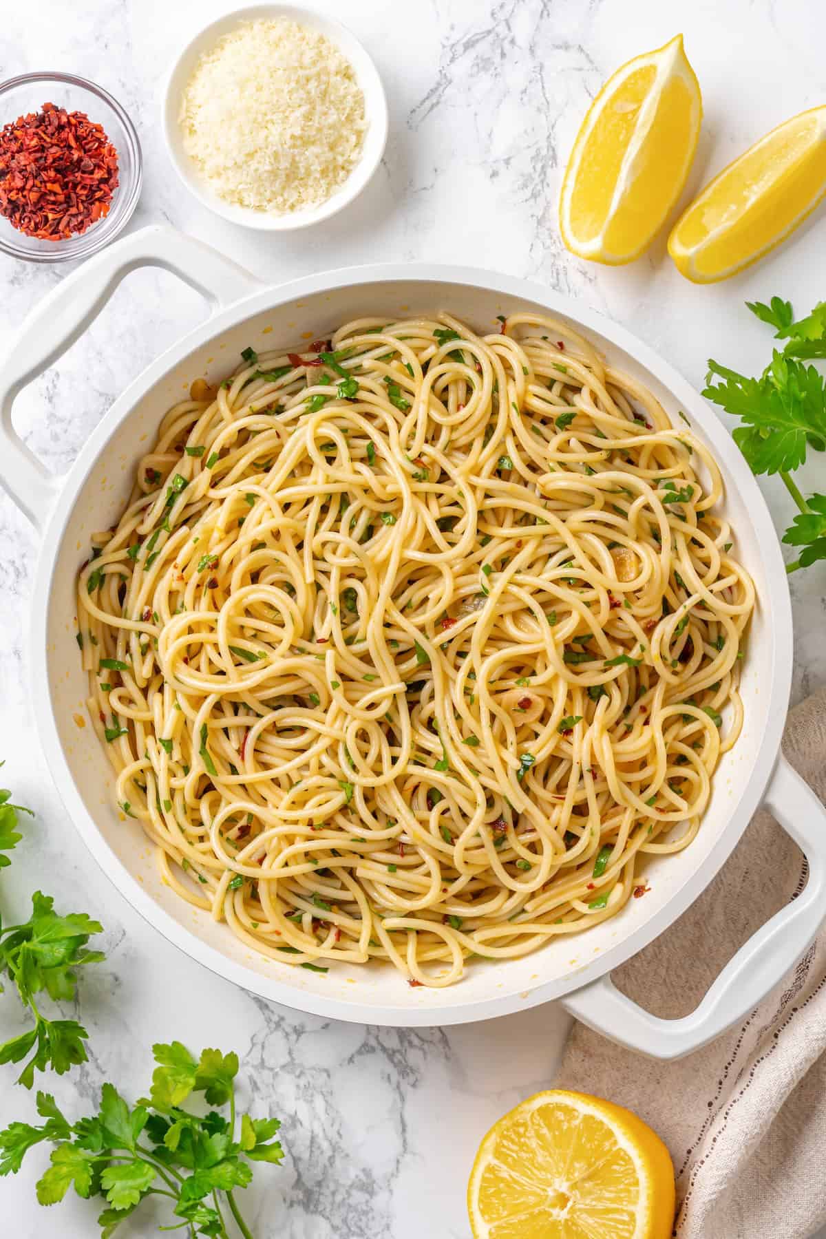 This easy aglio e olio features al dente spaghetti coated in a velvety garlic-infused sauce with a touch of heat from red pepper flakes. Aglio e olio in a pan surrounded by lemon wedges, red pepper flakes, vegan parmesan, and parsley.