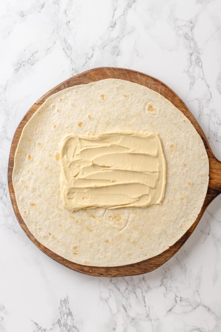 These 20 minute no-cook veggie wraps are packed with creamy hummus and avocado, crisp veggies, bright lime, and a drizzle of peanut sauce. A tortilla with hummus spread in the center.