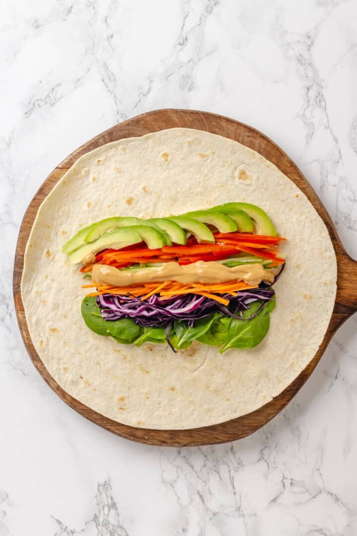These 20 minute no-cook veggie wraps are packed with creamy hummus and avocado, crisp veggies, bright lime, and a drizzle of peanut sauce. Red cabbage, carrot, cucumber, red pepper, avocado, spinach, and Thai peanut sauce, layered over hummus on a tortilla.