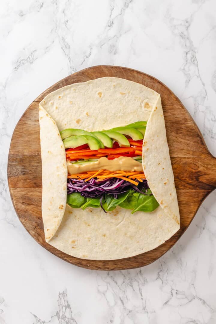 These 20 minute no-cook veggie wraps are packed with creamy hummus and avocado, crisp veggies, bright lime, and a drizzle of peanut sauce. Folding the sides of a tortilla over crunchy veggies, hummus, spinach, avocado, and peanut sauce.