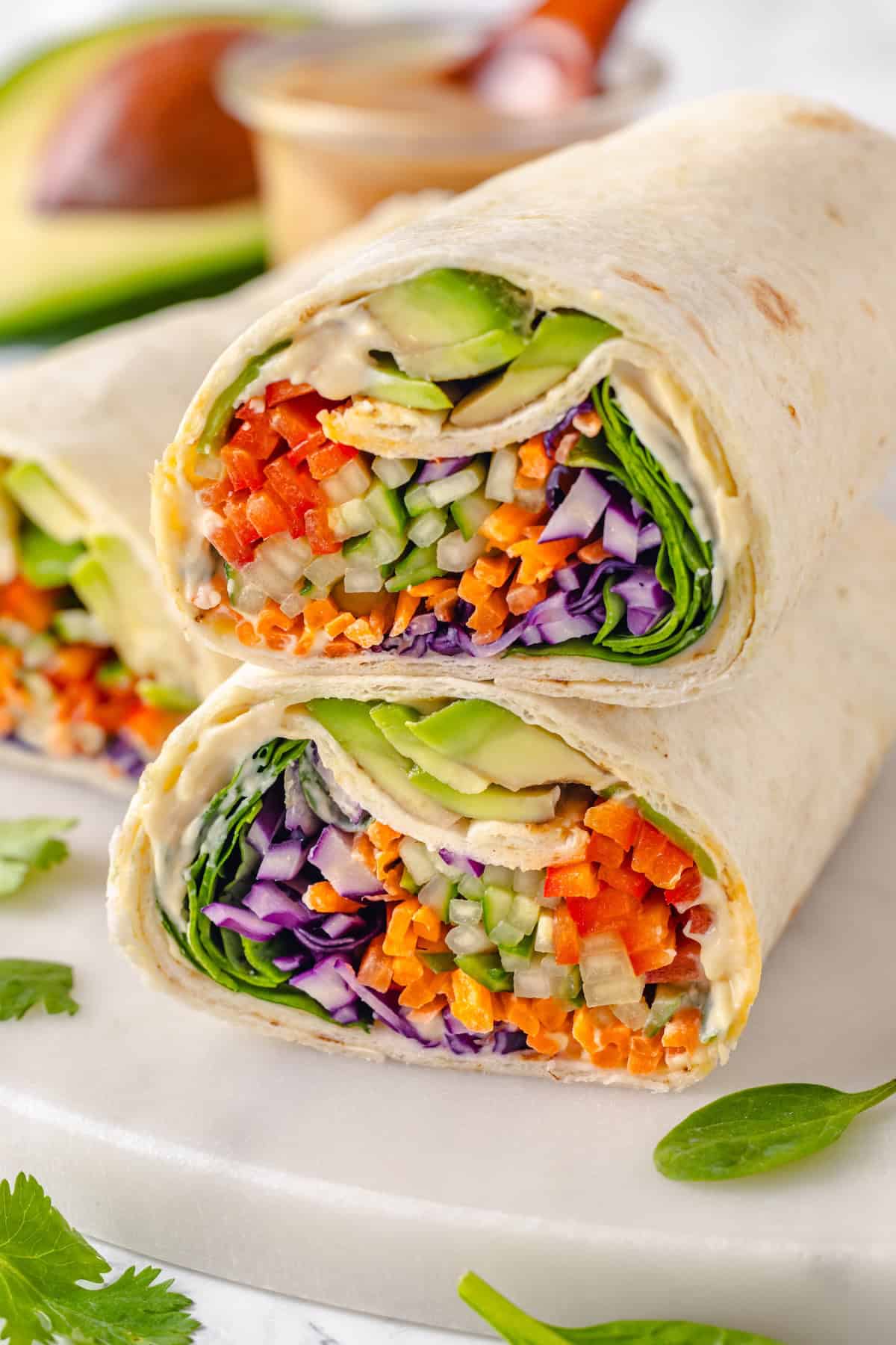 These 20 minute no-cook veggie wraps are packed with creamy hummus and avocado, crisp veggies, bright lime, and a drizzle of peanut sauce. A hummus veggie wrap cut in half to reveal the inside.