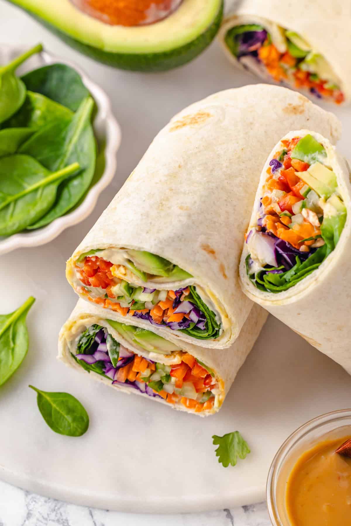 These 20 minute no-cook veggie wraps are packed with creamy hummus and avocado, crisp veggies, bright lime, and a drizzle of peanut sauce. Close-up of veggie wraps stacked on top of each other.
