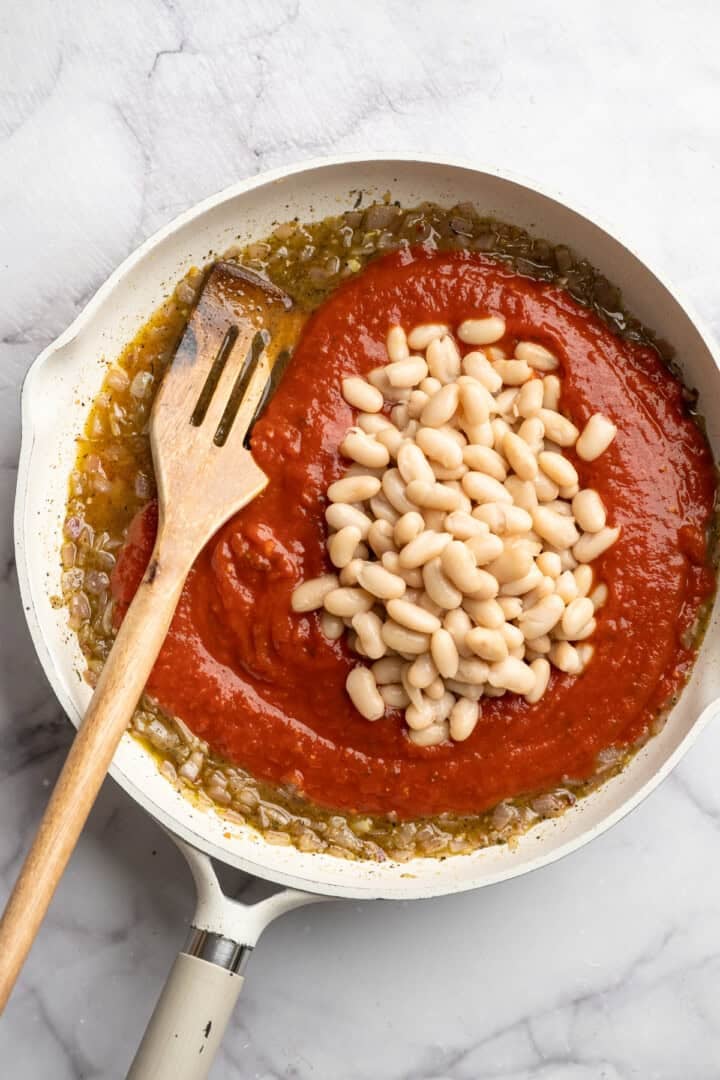 These 30-minute smoky white beans in tomato sauce are made with liquid smoke and plenty of garlic. They're amazing on toast for breakfast! Adding tomato sauce and white beans to a skillets with sautéed onion and garlic in white wine.