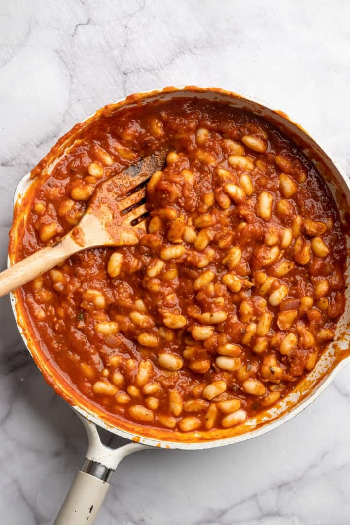 These 30-minute smoky white beans in tomato sauce are made with liquid smoke and plenty of garlic. They're amazing on toast for breakfast! Stirring white beans in tomato sauce in a skillet.