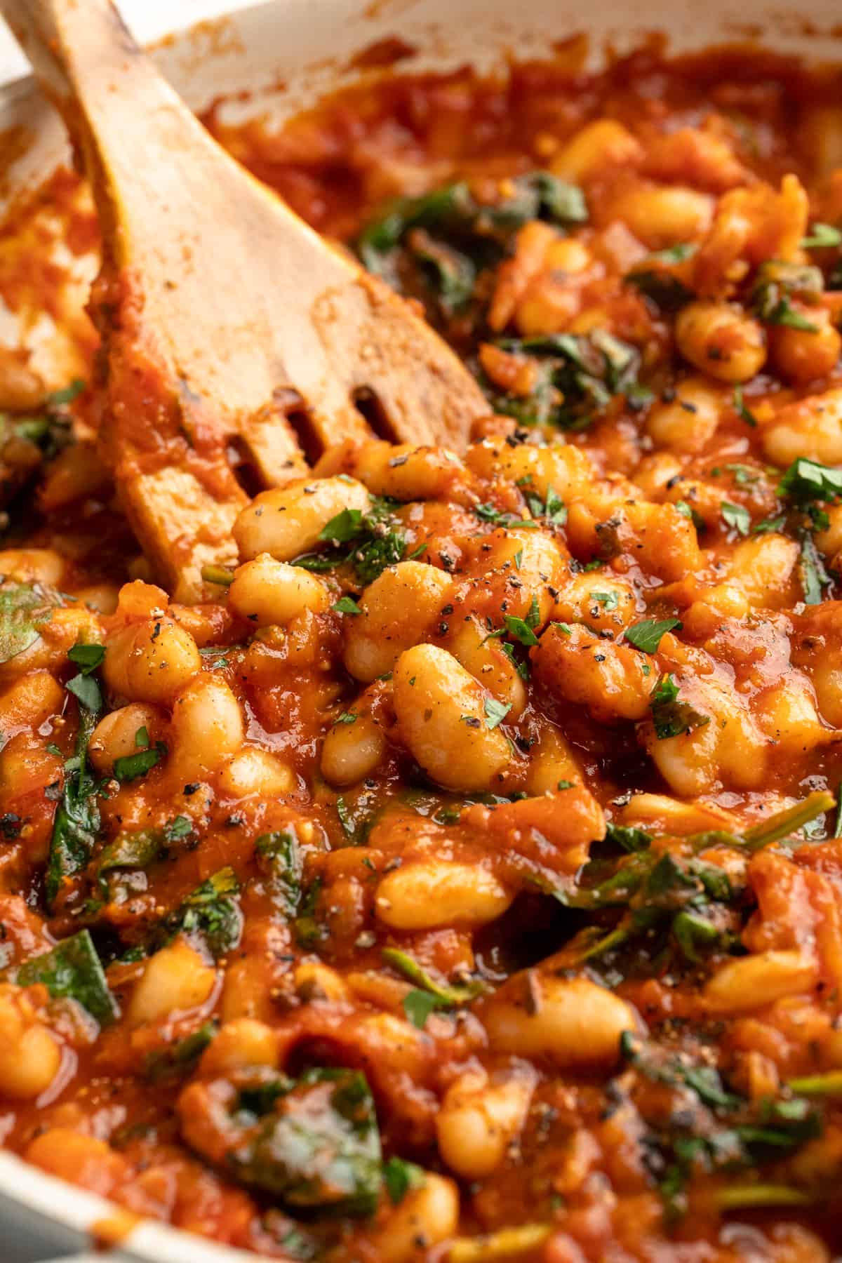 These 30-minute smoky white beans in tomato sauce are made with liquid smoke and plenty of garlic. They're amazing on toast for breakfast! Close-up of a wooden spatula stirring smoky white beans in tomato sauce.