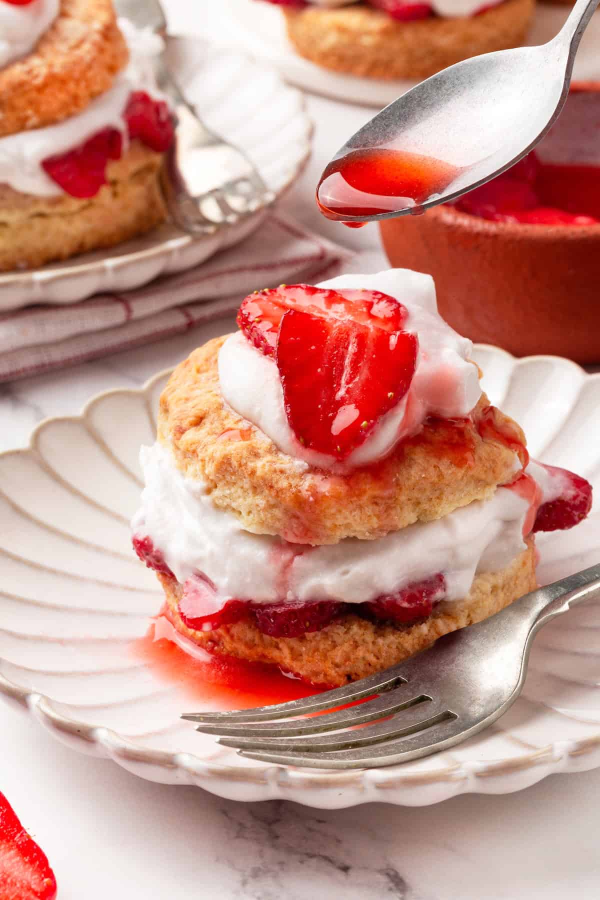 Strawberry shortcake recipe sandwiches sweet, syrupy macerated strawberries and fluffy vegan whipped cream between flaky homemade biscuits. A spoon drizzling syrup from macerated strawberries over a serving of strawberry shortcake.