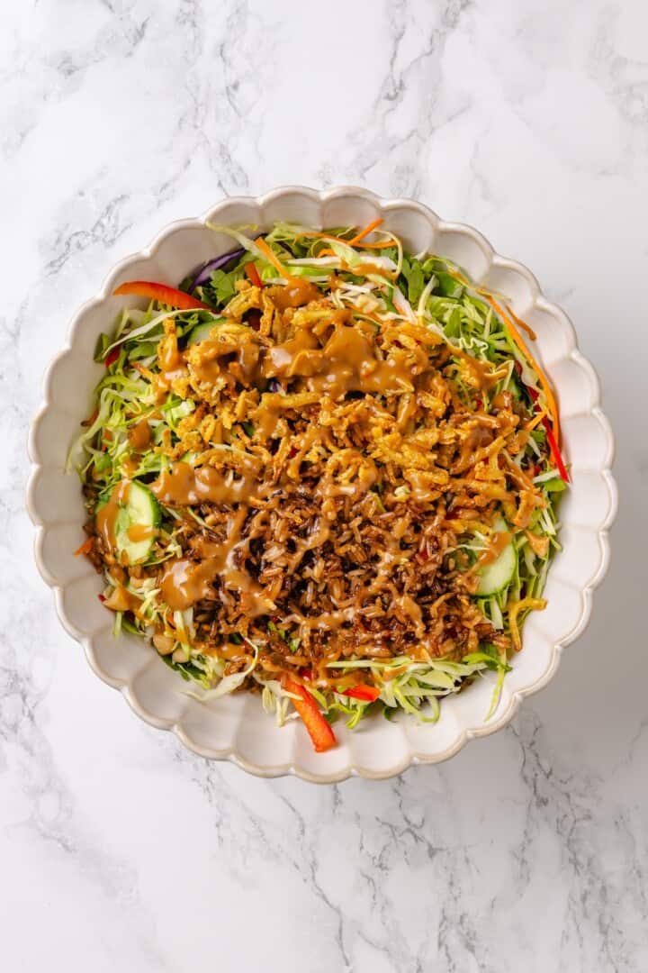 This crispy rice salad is made with soy-infused baked rice tossed with crunchy veggies, caramelized roasted tofu, and Thai peanut sauce. Adding crispy rice, baked tofu, and peanut sauce to a salad bowl loaded with crunchy veggies.