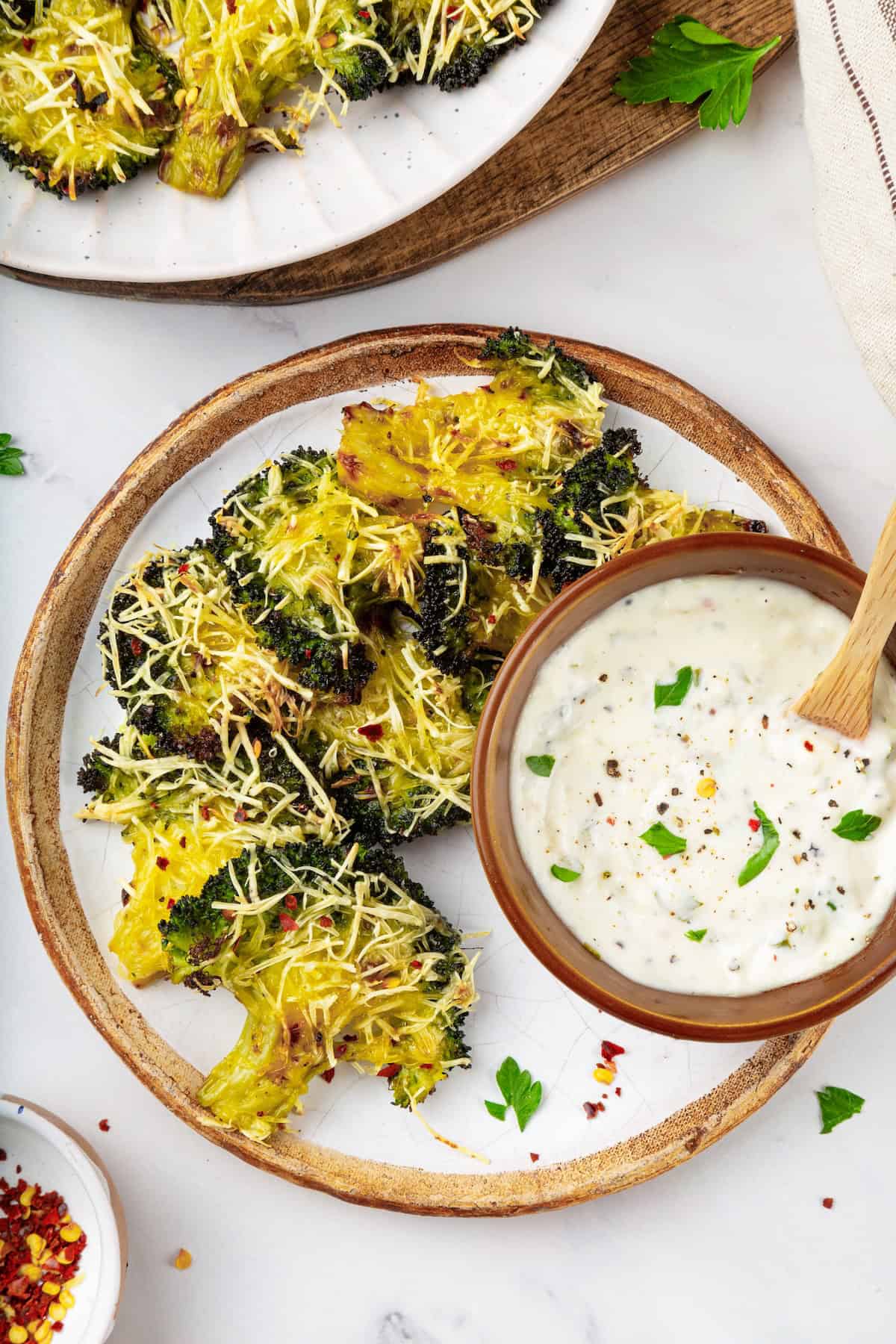 Cheesy vegan smashed broccoli is the perfect way to mix up your veggie routine. Served with a creamy garlic sauce, it's sure to impress. A plate of smashed broccoli with a bowl of vegan creamy garlic sauce.