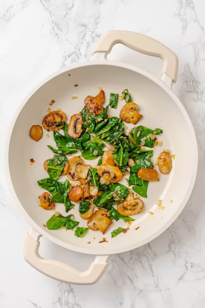 Start today right with this vegan omelette! Chickpea flour and water combine to make a tender omelette filled with veggies and vegan cheese. A pan with wilted spinach and sautéed mushrooms and shallot.