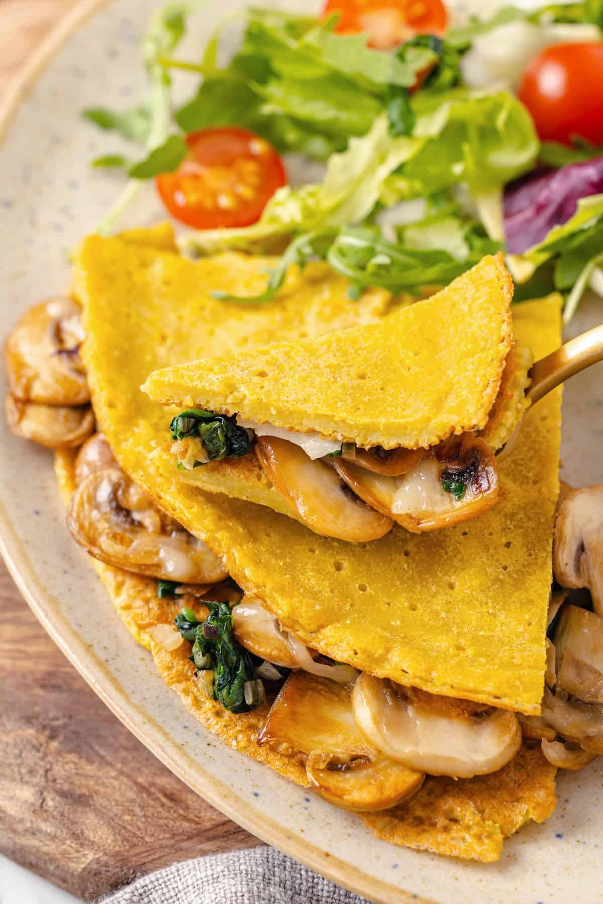 Start today right with this vegan omelette! Chickpea flour and water combine to make a tender omelette filled with veggies and vegan cheese. Close-up image of a fork picking up a bite of vegan chickpea flour omelette.