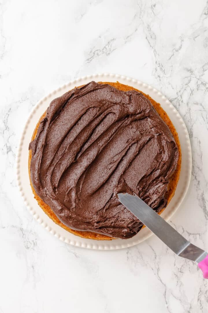 This vegan yellow cake is made of two layers of moist, tender, golden-yellow cake, filled and frosted with silky chocolate buttercream. Frosting the bottom cake layer with chocolate frosting.