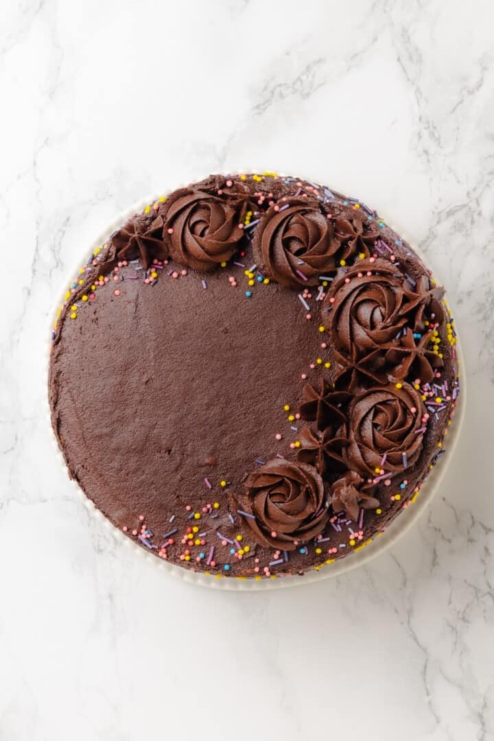 This vegan yellow cake is made of two layers of moist, tender, golden-yellow cake, filled and frosted with silky chocolate buttercream. Overhead image of vegan yellow cake with chocolate frosting and rainbow sprinkles.