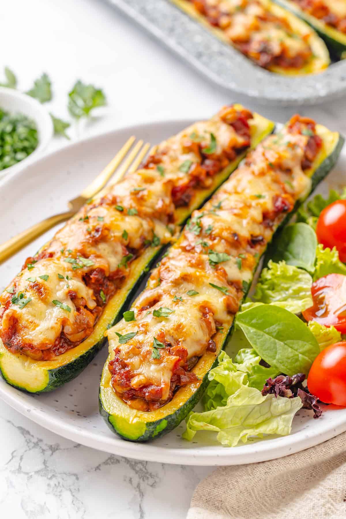 These stuffed zucchini boats are loaded with rich marinara sauce, melty vegan mozzarella, and protein-packed lentils. The perfect summer side. Two lentil-stuffed zucchini boats on a plate. with a green salad.