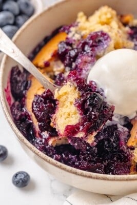 A spoon scooping into a serving of blueberry cobbler with vanilla ice cream.