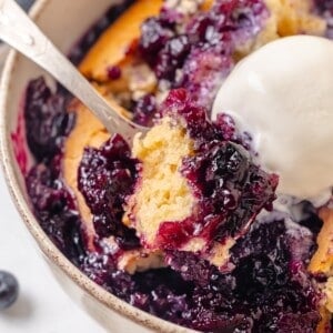 A spoon scooping into a serving of blueberry cobbler with vanilla ice cream.