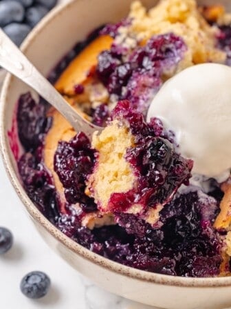 A spoon scooping into a serving of blueberry cobbler with vanilla ice cream.