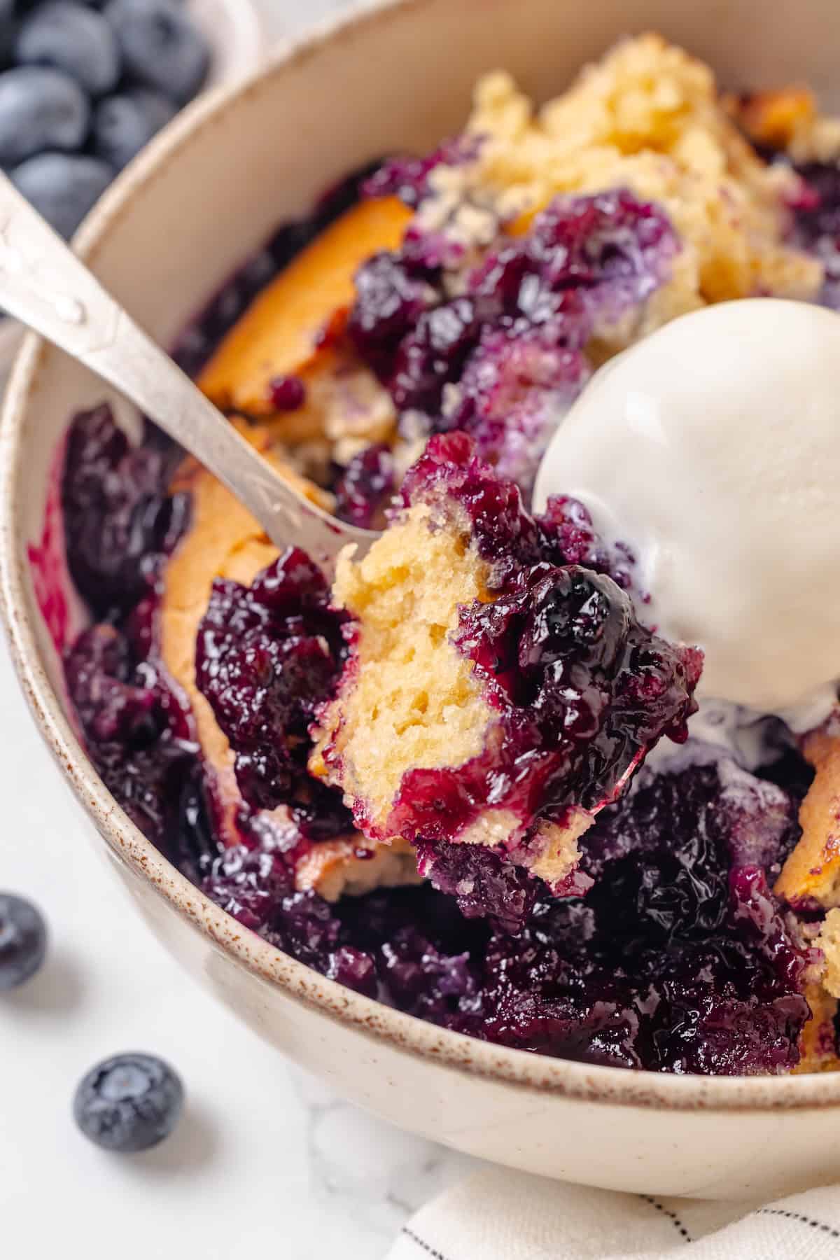 A spoon scooping into a serving of blueberry cobbler with vanilla ice cream.
