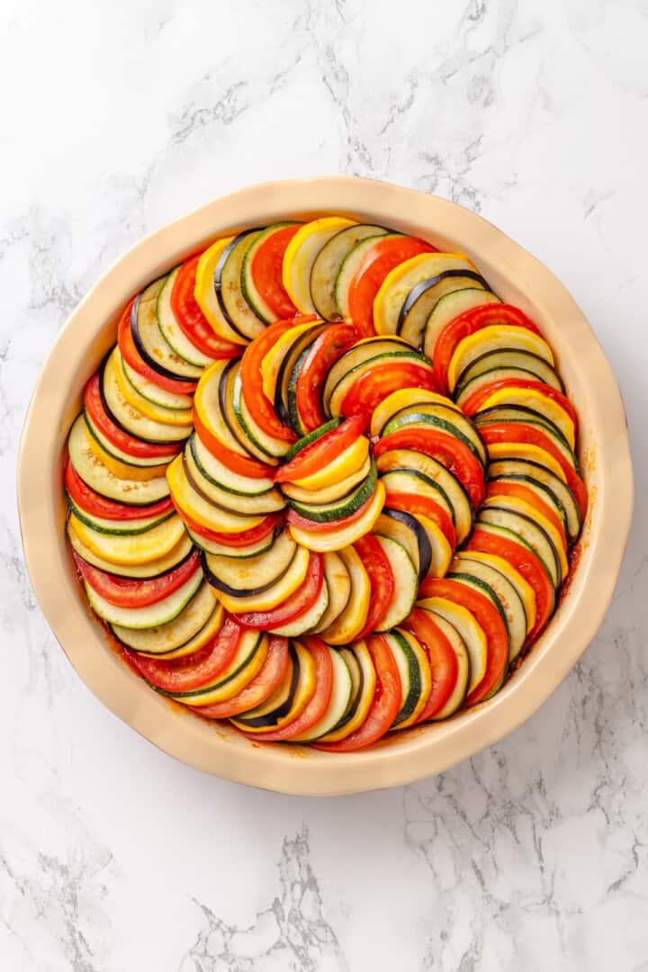 This easy ratatouille recipe boasts colourful layers of eggplant, squash, and tomato in a rich, tangy tomato sauce with delicate herb oil. A spiral of eggplant, zucchini, yellow squash, and tomatoes set over a layer of tomato sauce in a baking dish.
