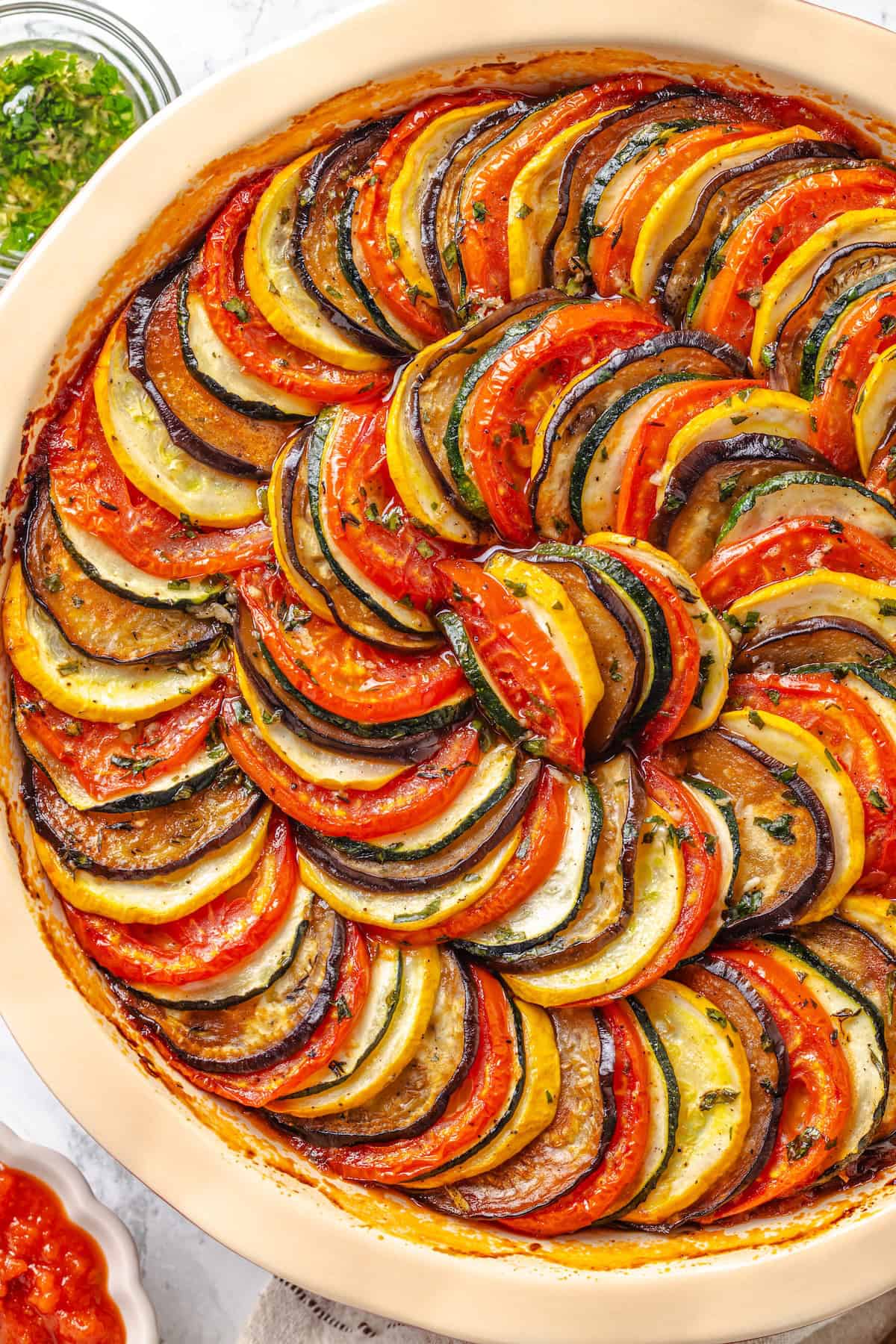 This easy ratatouille recipe boasts colourful layers of eggplant, squash, and tomato in a rich, tangy tomato sauce with delicate herb oil. Overhead image of a baking dish full of easy French ratatouille.
