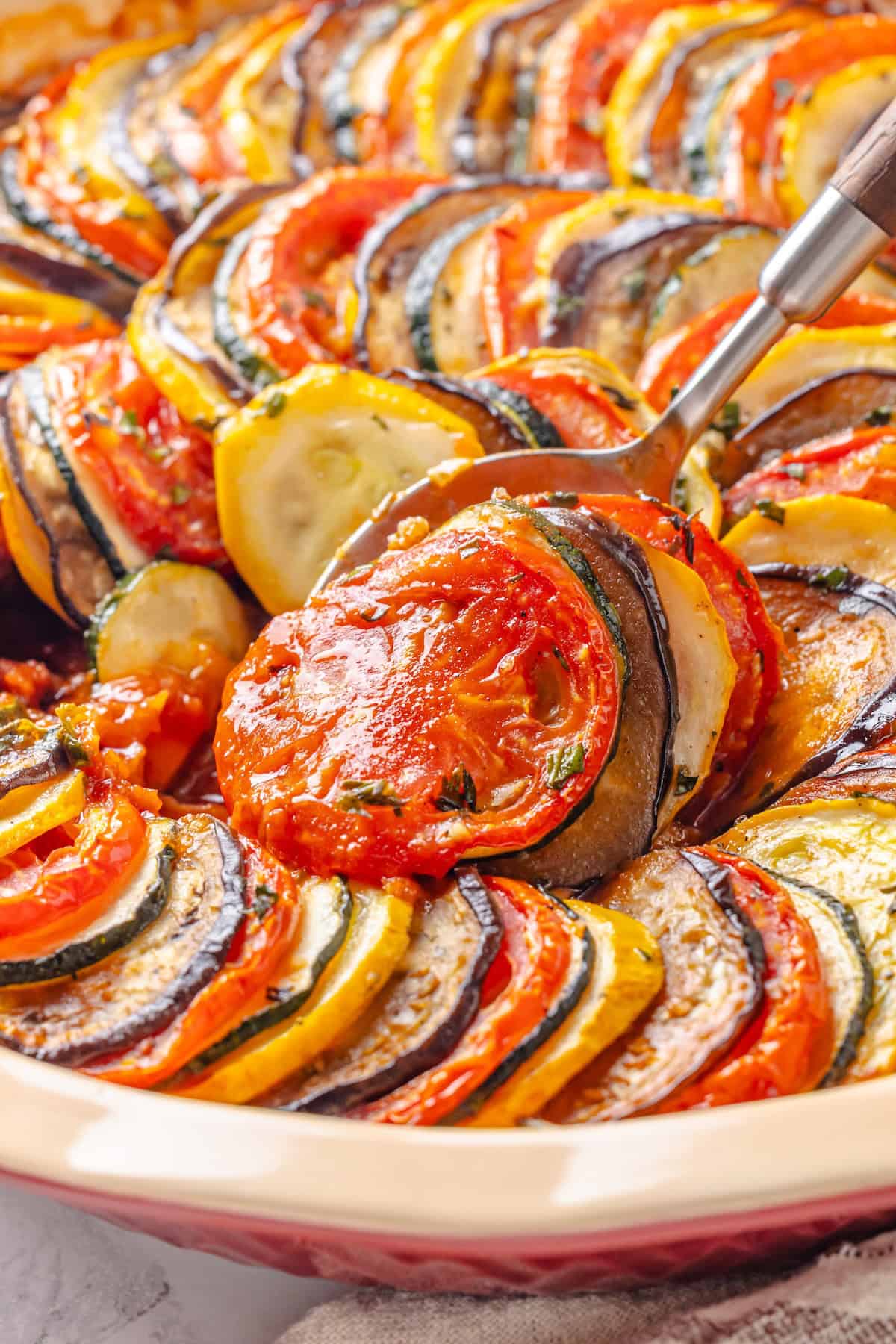 This easy ratatouille recipe boasts colourful layers of eggplant, squash, and tomato in a rich, tangy tomato sauce with delicate herb oil. A spoon diving into a baking dish of classic French ratatouille.