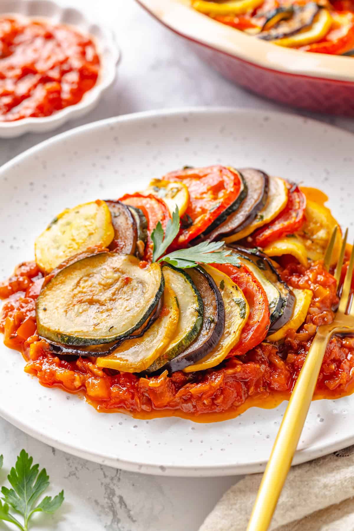 This easy ratatouille recipe boasts colourful layers of eggplant, squash, and tomato in a rich, tangy tomato sauce with delicate herb oil. A serving of classic French ratatouille on a plate with a gold fork.