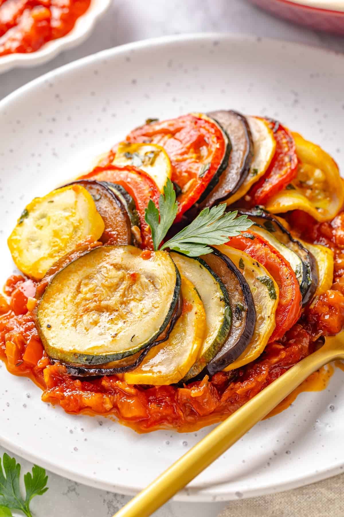 This easy ratatouille recipe boasts colourful layers of eggplant, squash, and tomato in a rich, tangy tomato sauce with delicate herb oil. A serving of ratatouille on a plate.
