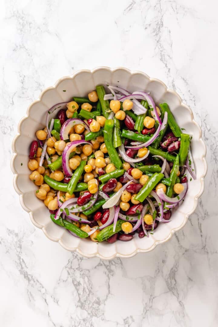 Kidney beans, chickpeas, blanched green beans, slivered red onion, and chopped fresh parsley in a salad bowl. Three bean salad tossed with maple-dijon apple cider vinegar dressing in a salad bowl.