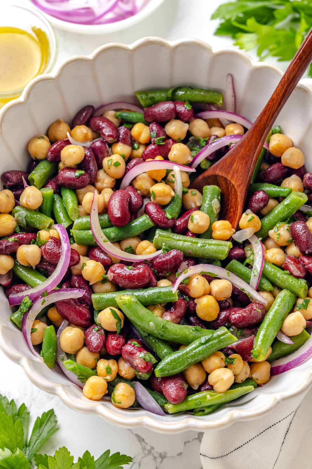 Kidney beans, chickpeas, blanched green beans, slivered red onion, and chopped fresh parsley in a salad bowl. Three bean salad in a serving bowl with a wooden spoon.