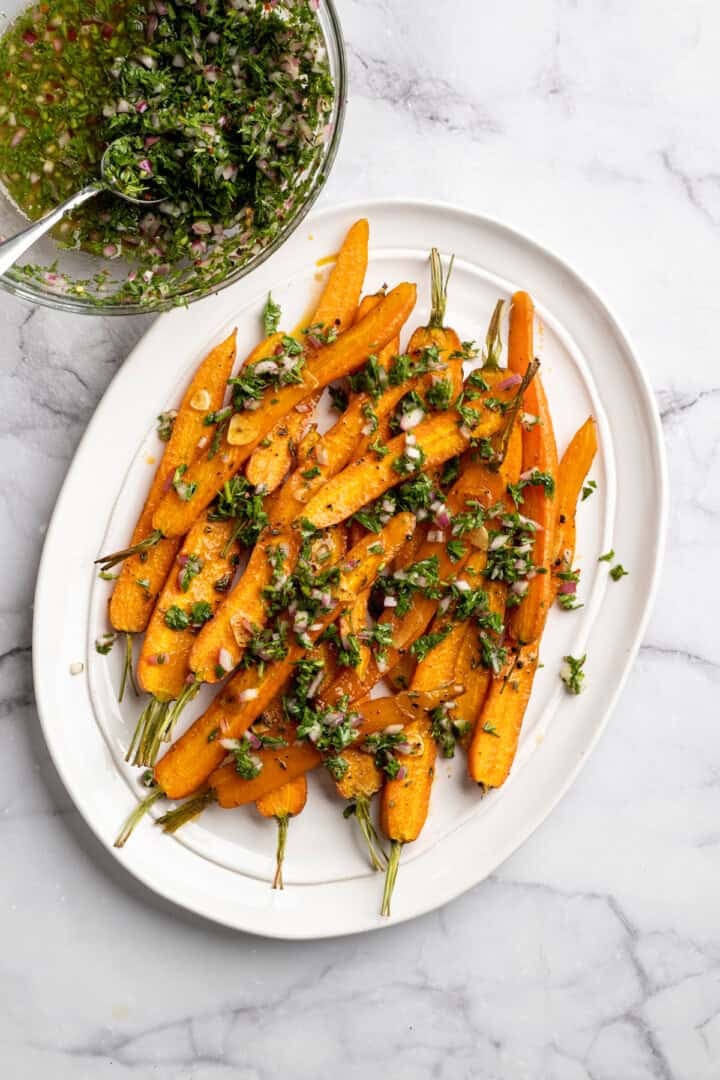 These perfectly caramelized maple roasted carrots are served with a vibrant chimichurri made from the carrot greens themselves. A plate of maple roasted carrots next to a bowl of carrot top chimichurri.